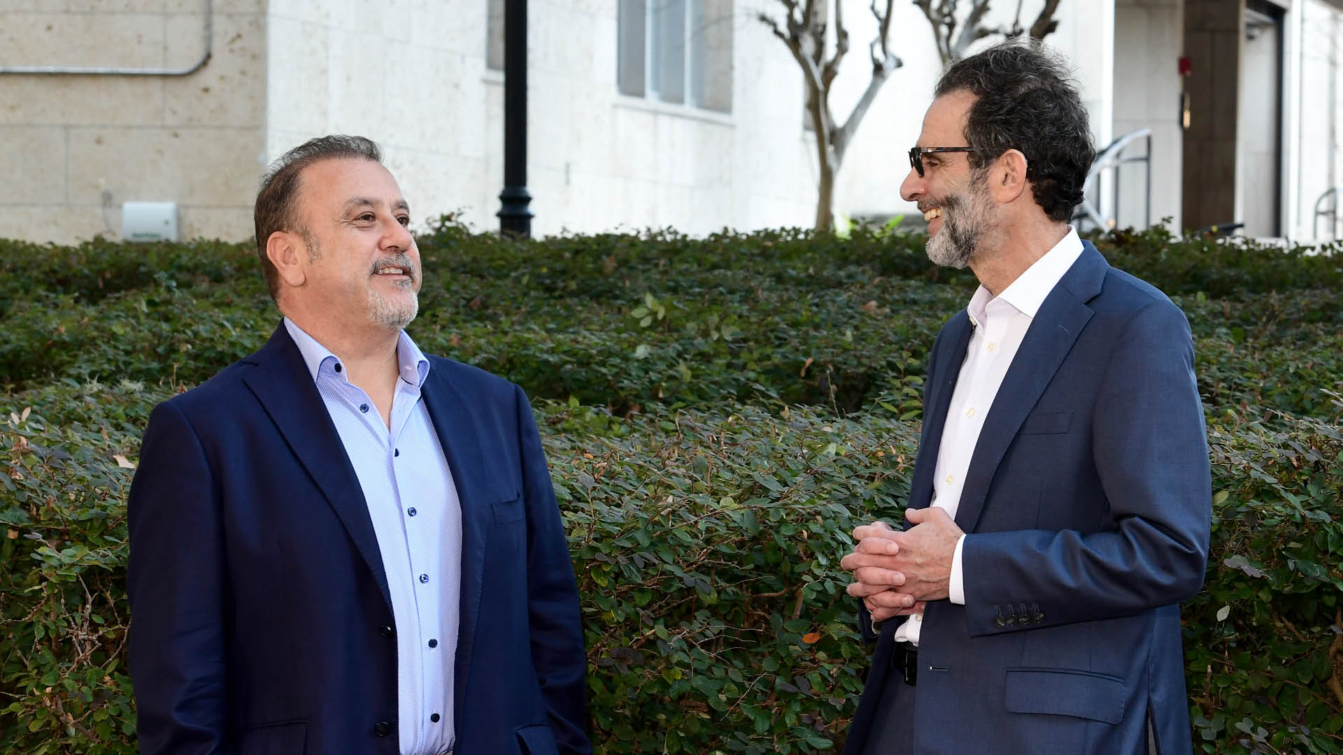 Two men in suits stand outdoors and talk near a hedge. One is wearing a light blue shirt and the other is wearing glasses and a white shirt. A building, trees, and a lamppost are in the background